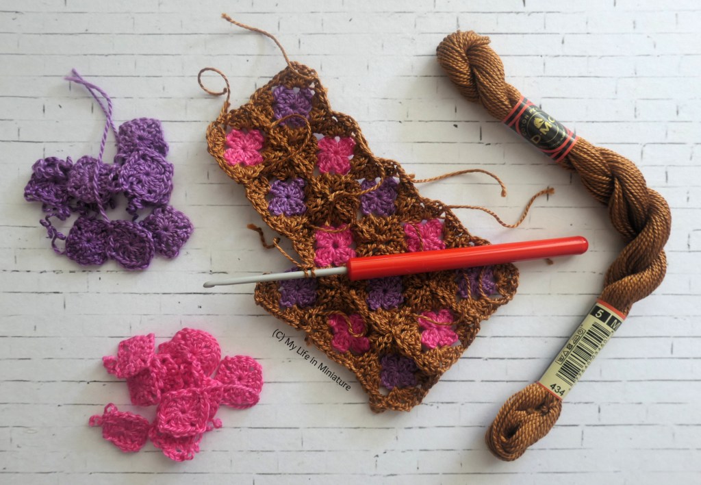A partially-completed miniature crochet blanket is photographed from above on a white brick background. A square in the third row is in the process of being attached. To the left of the blanket are, again, two piles of crochet squares, one pink and one purple. To the right is a new skein of copper yarn.