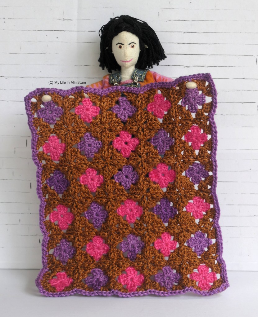 Tiffany stands in front of a white brick background, holding a rectangular crocheted blanket in front of her. The blanket is made of granny squares with alternating pink and purple centres, surrounded by copper brown. The border of the blanket is purple. 