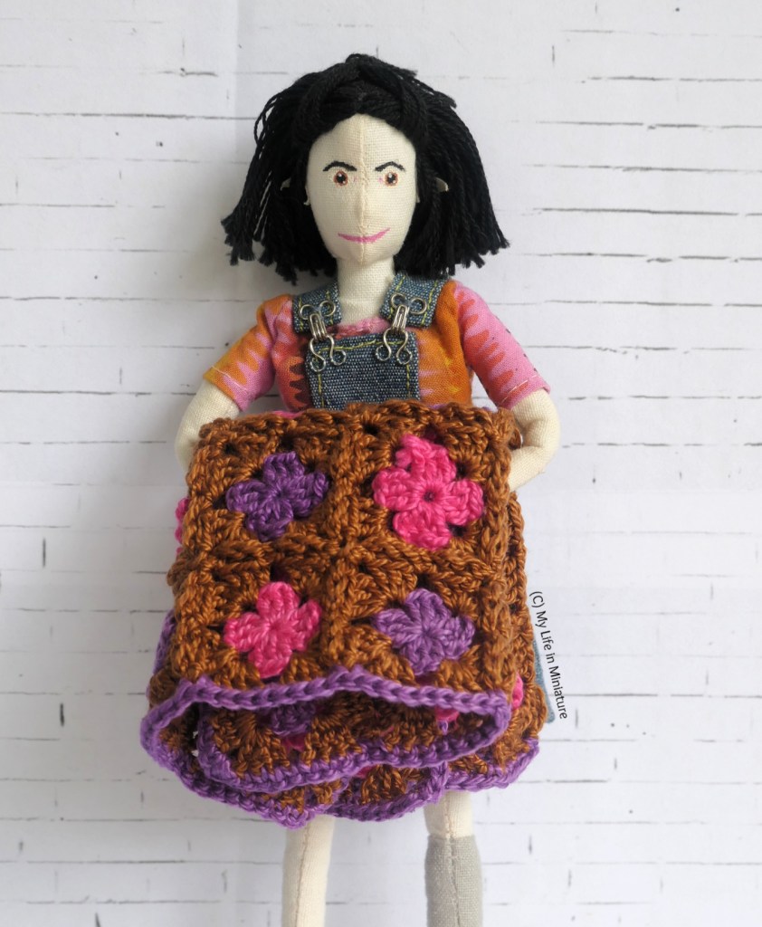 Tiffany stands in front of a white beick background, again holding the crocheted blanket in front of her. This time, however, it is folded up and draped over her arms. She's wearing a pink and orange shirt under denim dungarees. 