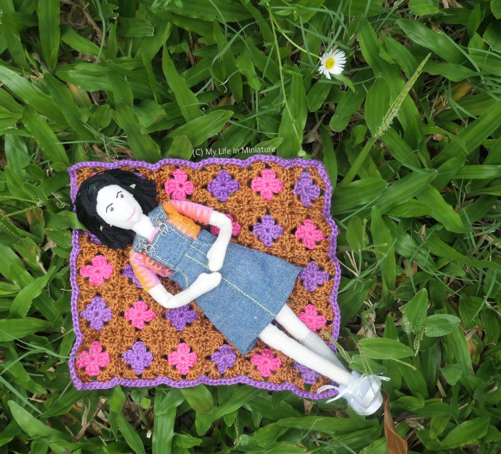 Tiffany is photographed from above, lying on her back on the crocheted blanket on grass. She's lying diagonally, so most of her is on the blanket. Her feet are crossed, her hands are on her stomach, and she looks relaxed. 