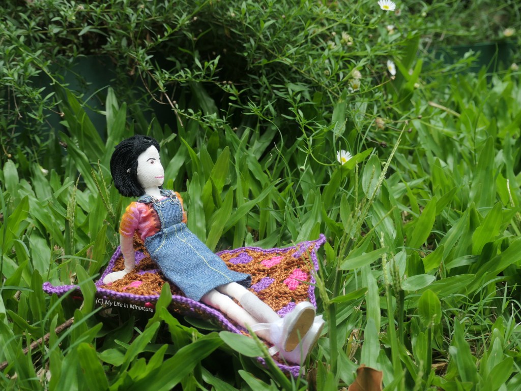 Tiffany sits on the crocheted blanket with her legs stretched out in front of her, leaning back on her arms. She's looking towards the bush of daisies in the background, and grass surrounds the blanket. 