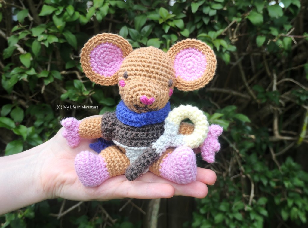 A crocheted mouse character, Lewt,  sits on the author's outstretched hand, with a hedge in the background. The mouse is mid-brown, with pink hands, feet, and inner ears. He is wearing a dark brown shirt, and a blue-purple cloak around his neck. He holds a short staff with a large gold ring at the top. 