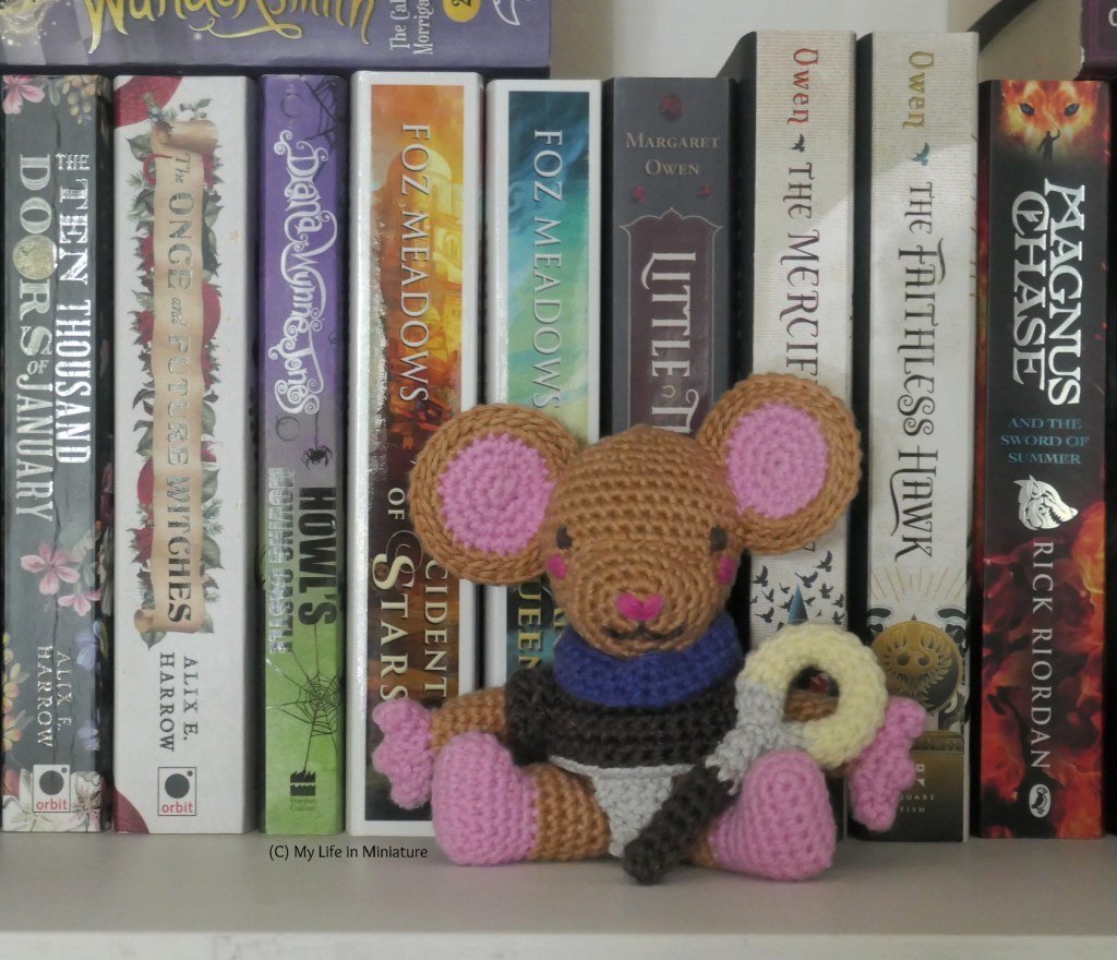 Lewt sits on the author's bookshelf, leaning against the spines of the books. He has cloak and staff, and faces the camera. The books behind him are a small selection of the author's fantasy novels. 