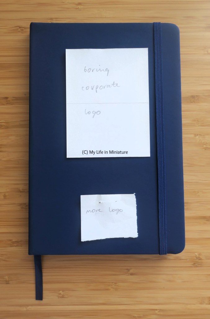 A navy blue notebook is photographed from above on a wood background. Two corporate logos on the cover are covered by scraps of paper that say "boring corporate logo" and similar on them. The notebook also has navy elastic holding it shut, and a navy ribbon page-marker.
