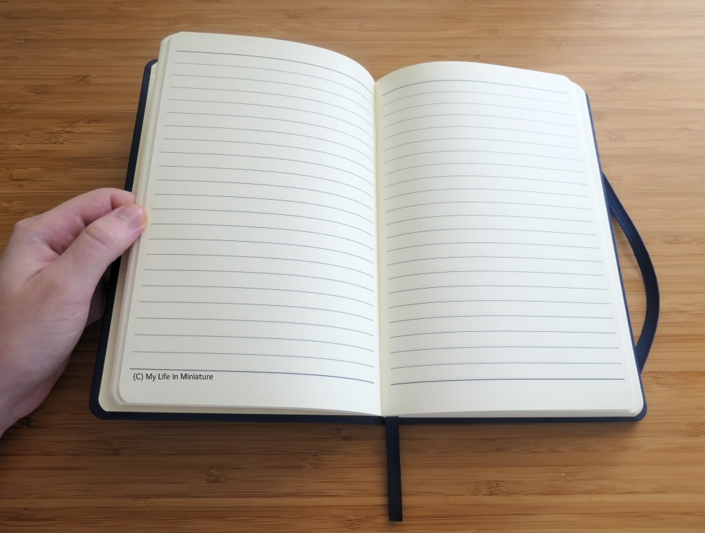 The author's hand holds the navy notebook open on a wood surface. The inside of the notebook is beige lined paper, with no corporate branding.