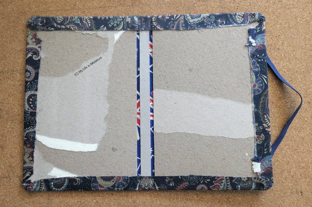 The cover of the notebook is photographed from above on a cork surface. The cardboard of the inside of it is visible, where the outer fabric has been wrapped around and the elastic glued in place.
