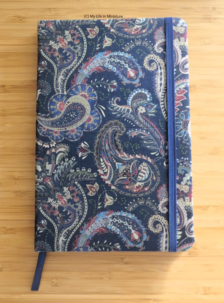 The completed notebook is photographed from above on a wood surface. The cover is facing up, which is now covered in the navy paisley cotton sateen fabric. The navy elsatic holds the noteobok shut, and the ribbon peeks out the bottom.