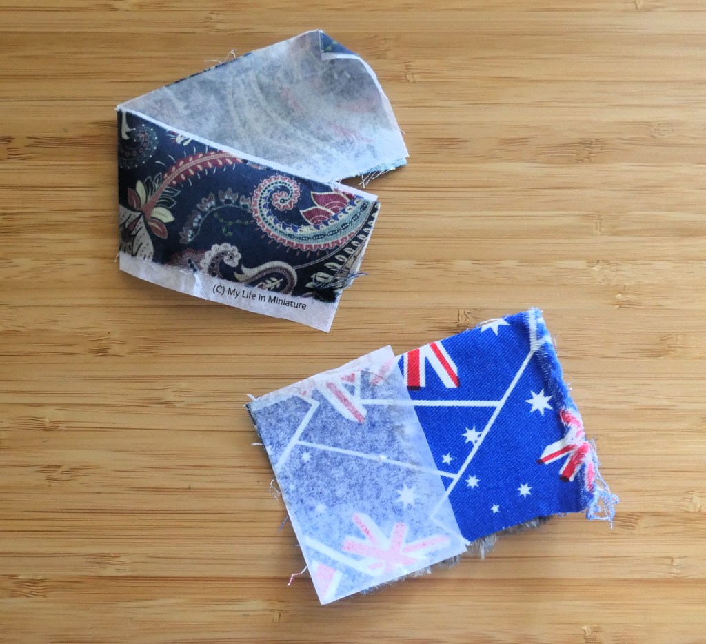 Two samples of fabric are photographed from above on a wood surface. The top one is a long strip of navy paisley cotton sateen, folded in half. The underside of the fabric has tissue paper glued to it. The bottom square is a twill fabric, half of which has tissue paper glued to it. The underside of the twill (which isn't visible) has the same navy cotton sateen glued to it.