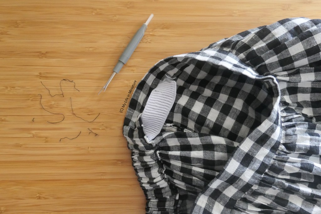 Close-up of the waistband of a black-and-white checked skirt, on a wood background. Part of said waistband has been unpicked to expose the elastic within. To the left of the skirt is a grey unpicker and some thread scraps.