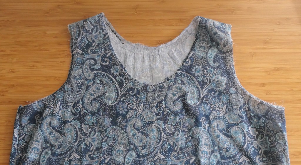 Close-up of the top half of a blue paisley-print tank top, on a wood background. The neck and armholes have their raw edges exposed.