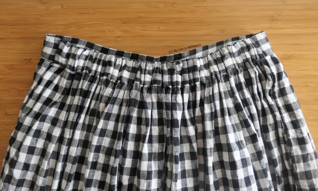 Close-up of the waistband of the black-and-white checked skirt, on a wood background. The waistband is fully assembled again, and the elastic in the back is shorter than the front flat section of waistband.
