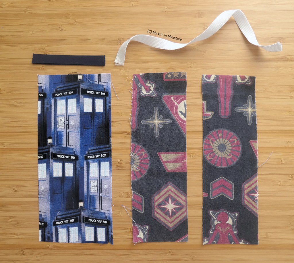 Three small rectangles of fabric are laid flat on a wood surface. They are all of similar size; one has blue TARDISes on it, and the other is grey and red. Above the rectangles is a small piece of navy bias binding and a small piece of elastic.