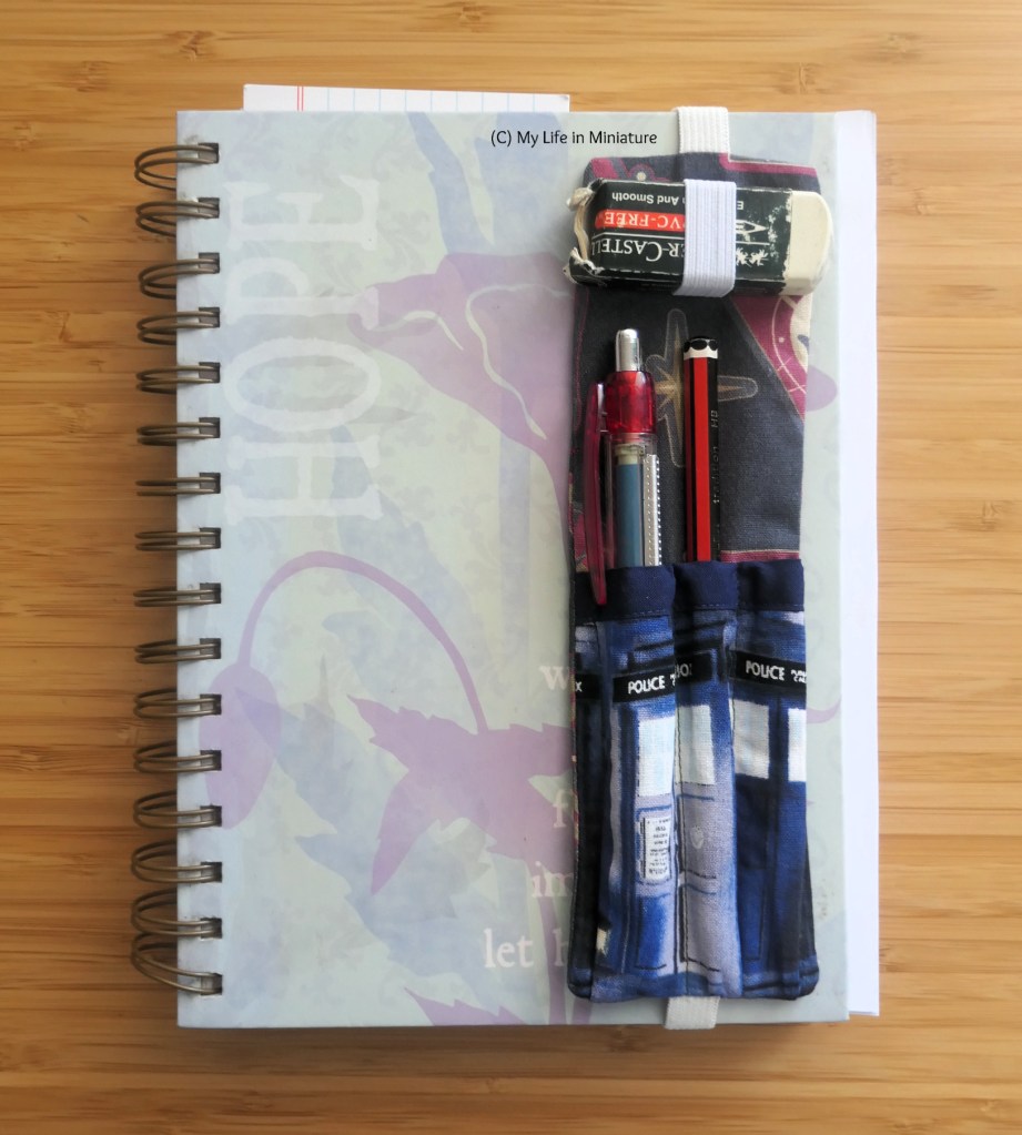 The pen holder is wrapped around an A5 notebook, with the elastic at the back and the pens at the front. Said notebook is laid flat on a wood surface. There are a couple of papers sticking out of the notebook.