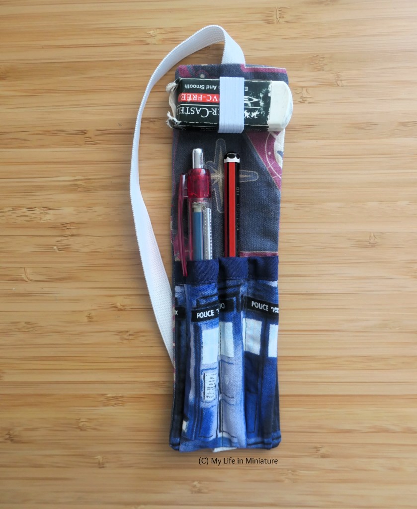 A fabric pen holder is laid flat on a wood surface. It is a rectangle of grey and red fabric, with elastic attached at each end to make a loop. There are three tubes sewn onto the bottom half with the TARDIS fabric, two of which hold writing implements. A loop of elastic at the top holds a rubber.