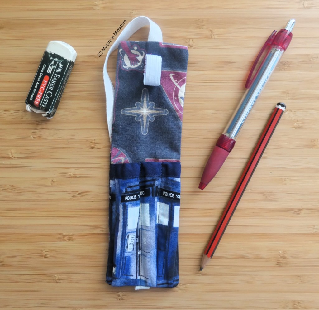 The pen holder is laid flat on a wood surface, without a notebook. It has been emptied of its contents, and the rubber, pen, and pencil are around it.