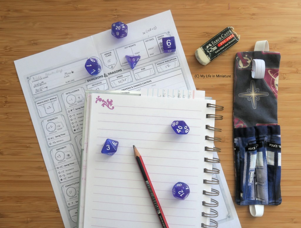The pen holder and the A5 notebook from earlier are laid flat on a wood surface. The notebook is open to a blank page, and is on top of a Dungeons and Dragons character sheet. The pen holder is beside the notebook, empty of the rubber and pencil. Scattered across the character sheet and notebook is a set of seven purple dice.