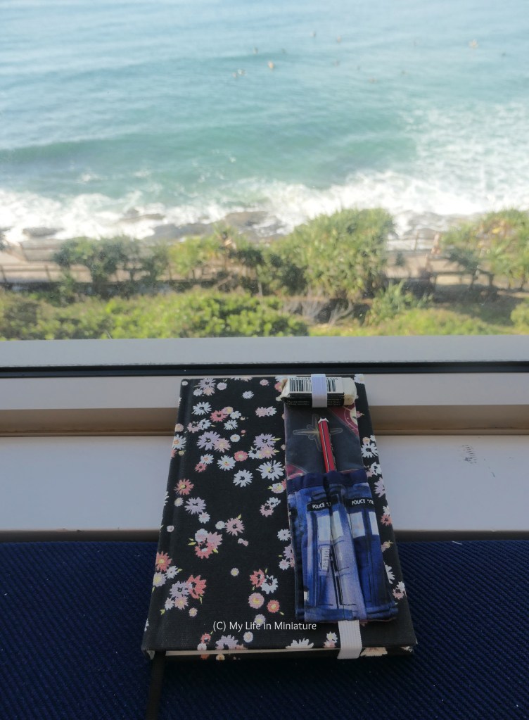 The pen holder is wrapped around a different A5 notebook, which is photographed on a windowsill. Outside the window is some trees, a boardwalk, and the sea. 
