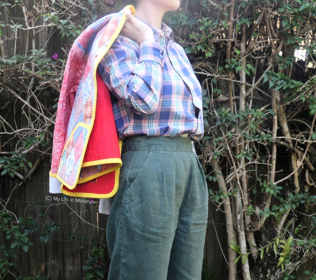 The author stands in front of a hedge, photographed from neck to thighs. She's almost side-on to the camera, with the quilted jacket thrown over the closer shoulder. She's wearing the checked shirt and green pants as well. 