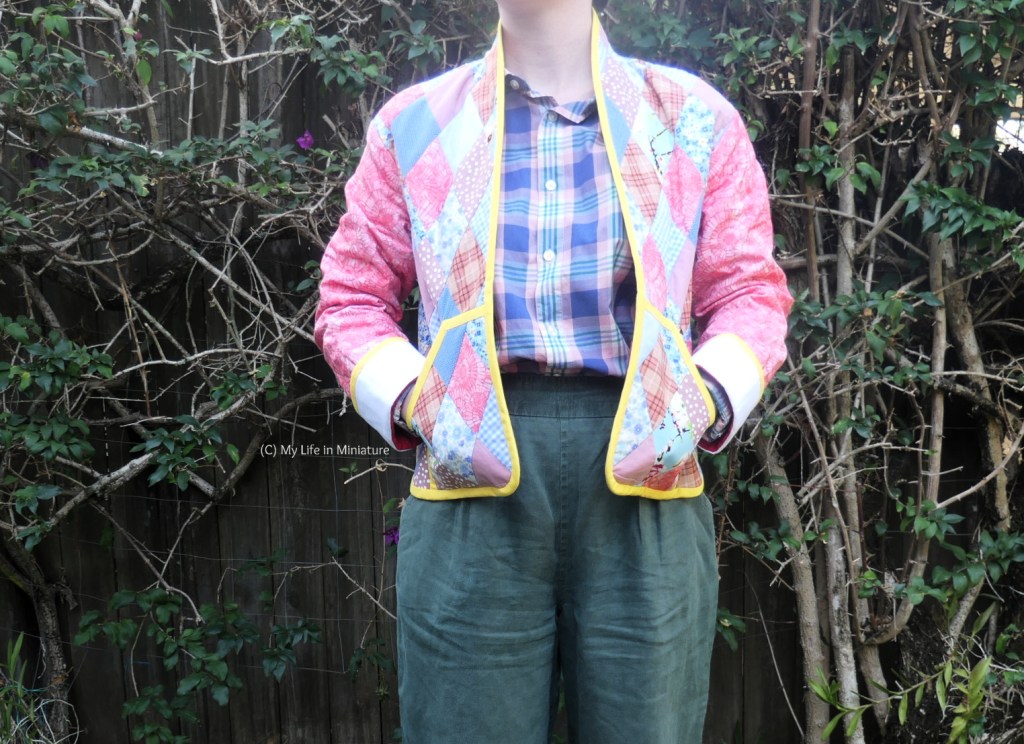 The author stands in front of a hedge, photographed from neck to thighs. She's wearing the quilted jaclet with her arms in her sleeves and her hands in the jacket's two front pockets. She also wears a blue checked shirt and green pants.