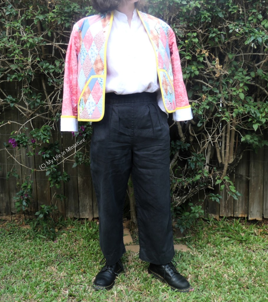 The author stands in front of a hedge, photographed from neck to the ground. She's wearing the quilted jacket over a white shirt, black pants, and black boots. The jacket is draped over her shoulders; her arms are not in the sleeves. Instead, her hands are tucked into her pockets underneath the jacket.