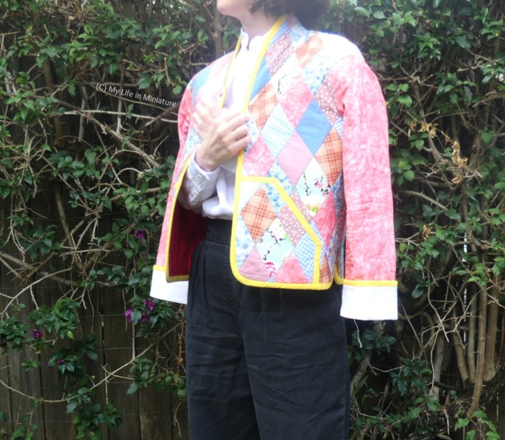 The author stands in front of a hedge, photographed from neck to knees. She is wearing a quilted jacket with a white shirt and black pants. The jacket bodice is patchworked with hundreds of small blue and pink diamonds of various patterns. The arms are plain pink, with white cuffs. Her body is at an angle to the camera, so one whole side of the front of the jacket is visible. 