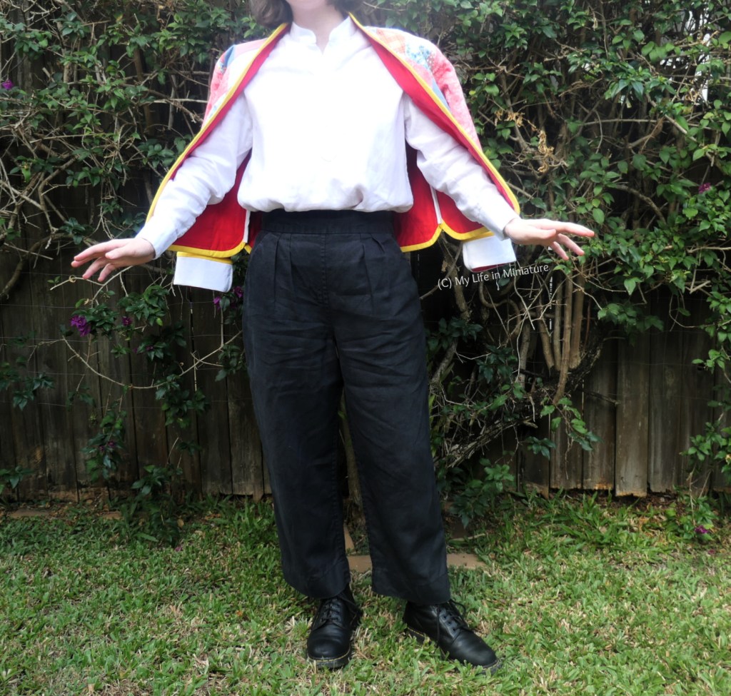 The author stands in front of a hedge, photographed from neck to the ground. She's wearing the quilted jacket over her shoulders, with her arns out of the sleeves, and a white shirt with black pants and boots. Her arms are out to her sides a little, with her fingers artfully spread. The red lining of the jacket is a little bit visible, as well as the patchworking at the shoulders. It looks very wizard-ish.