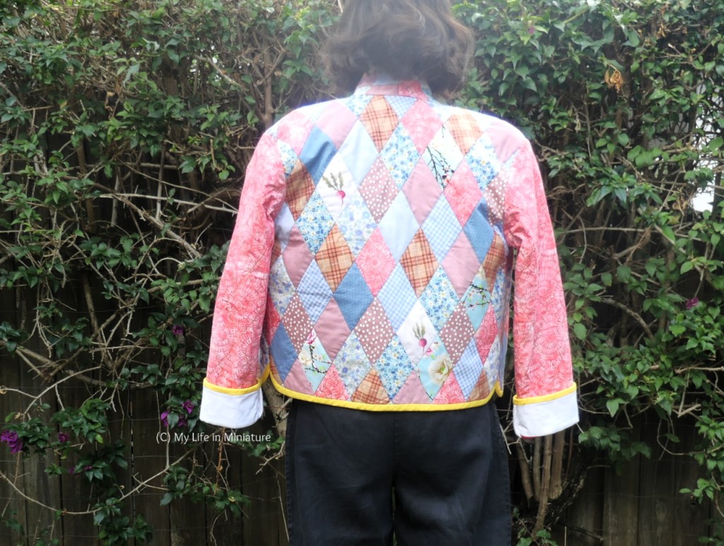 The author stands in front of a hedge, photographed from head to thighs. She's wearing the quilted jacket over her shoulders, and is facing away from the camera, so the back of the jacket is in view. It is completely patchworked with pale blue and pink fabrics in a variety of patterns. Her black pants are also visible beneath the jacket.
