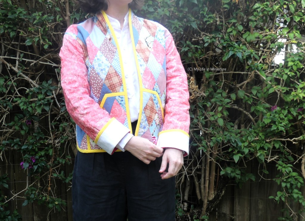 The author stands in front of a hedge, photographed from neck to thighs. She's wearing the quilted jacket properly, with a white shirt and black pants. One hand is tugging at the cuff of the other sleeve.