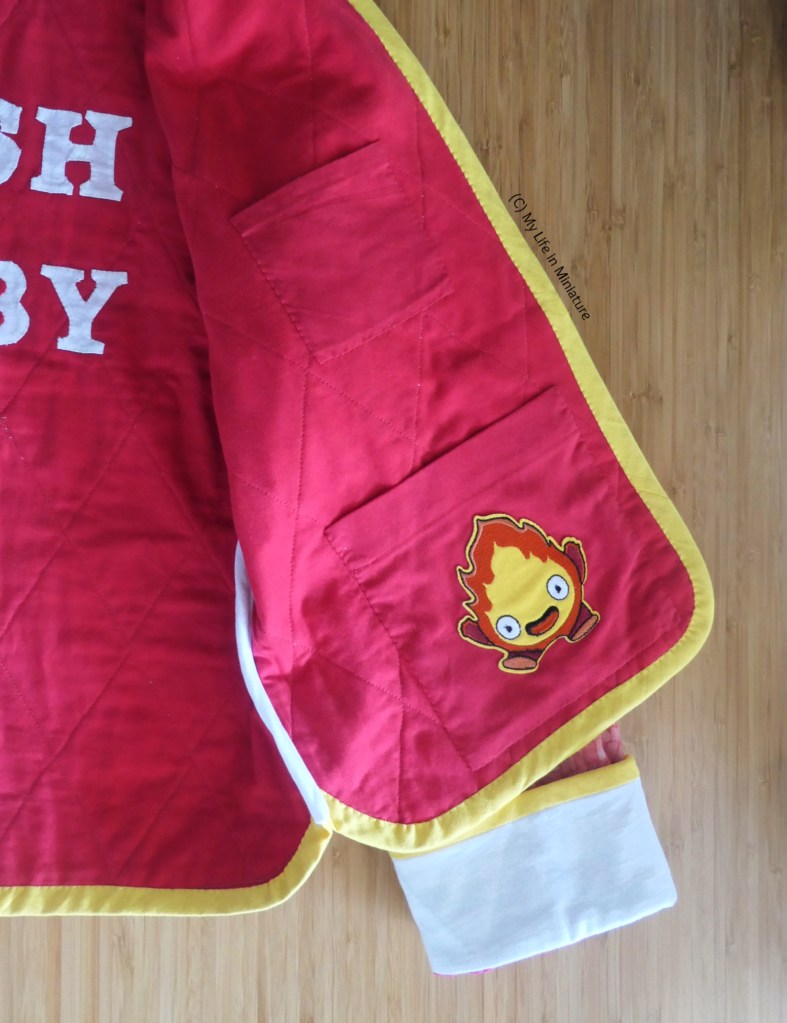 Close-up of the inside of one of the front panels of the jacket. There are two patch pockets hand-sewn onto the jacket: a large one near the bottom, and a smaller one further up. The bottom pocket has an iron-on patch of Calcifer the fire demon on it.