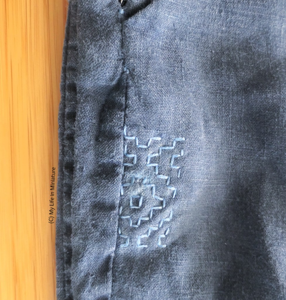 Close-up of the repair to the hole on the navy pants. The hole has been stitched over with small stitches in pale blue thread, to make a blocky star-like pattern.