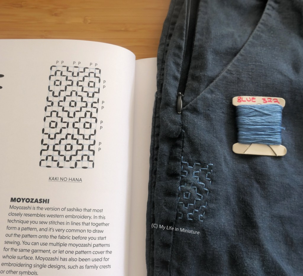 The sashiko area of the pants is laid across an open book. The opposing page of the book has a diagram of the pattern on the pants, called "Kaki no hana". The skein of blue thread used in the stitching is laid on the pants.