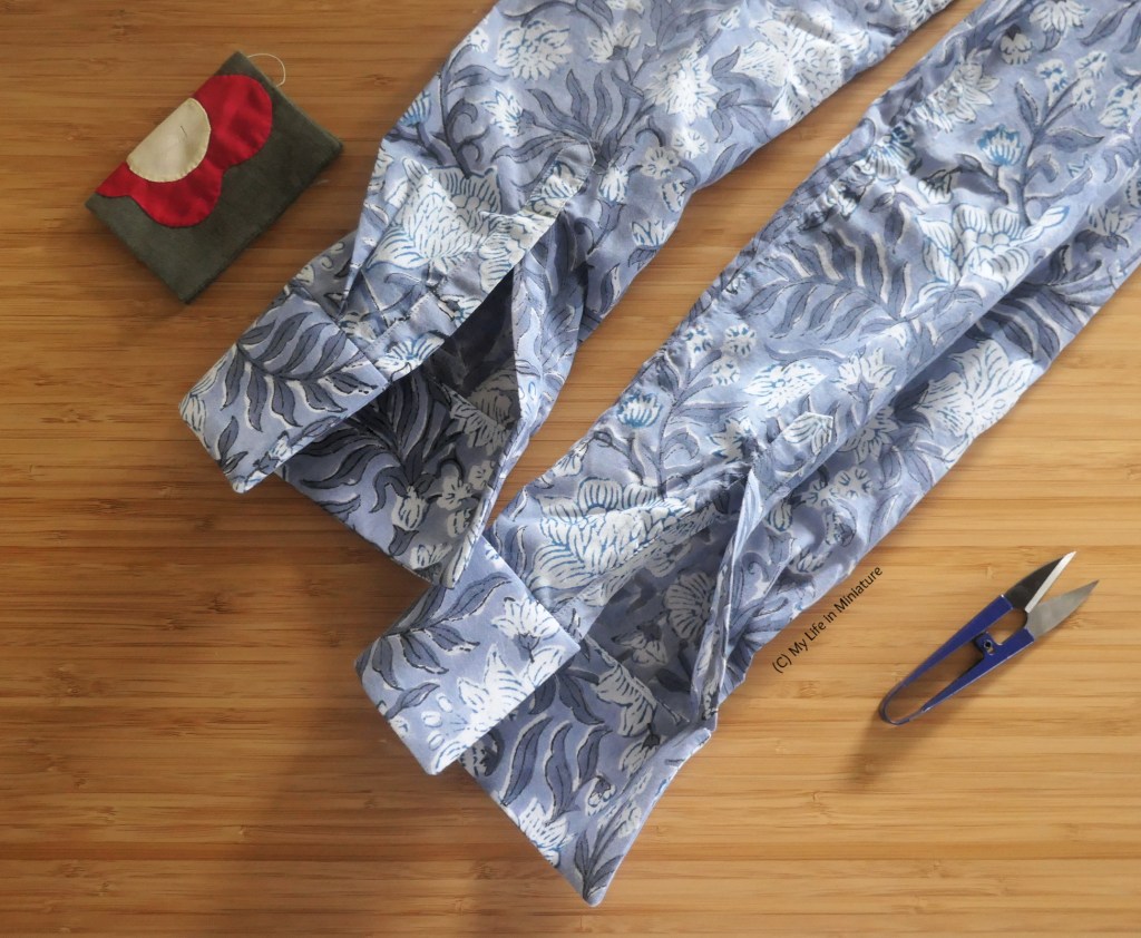 Two unattached shirt sleeves are laid flat on a wood surface, angling from bottom-left to top-right. They are made of grey cotton with a white and darker-grey floral design. Beside them is a needlebook and a pair of thread snips.