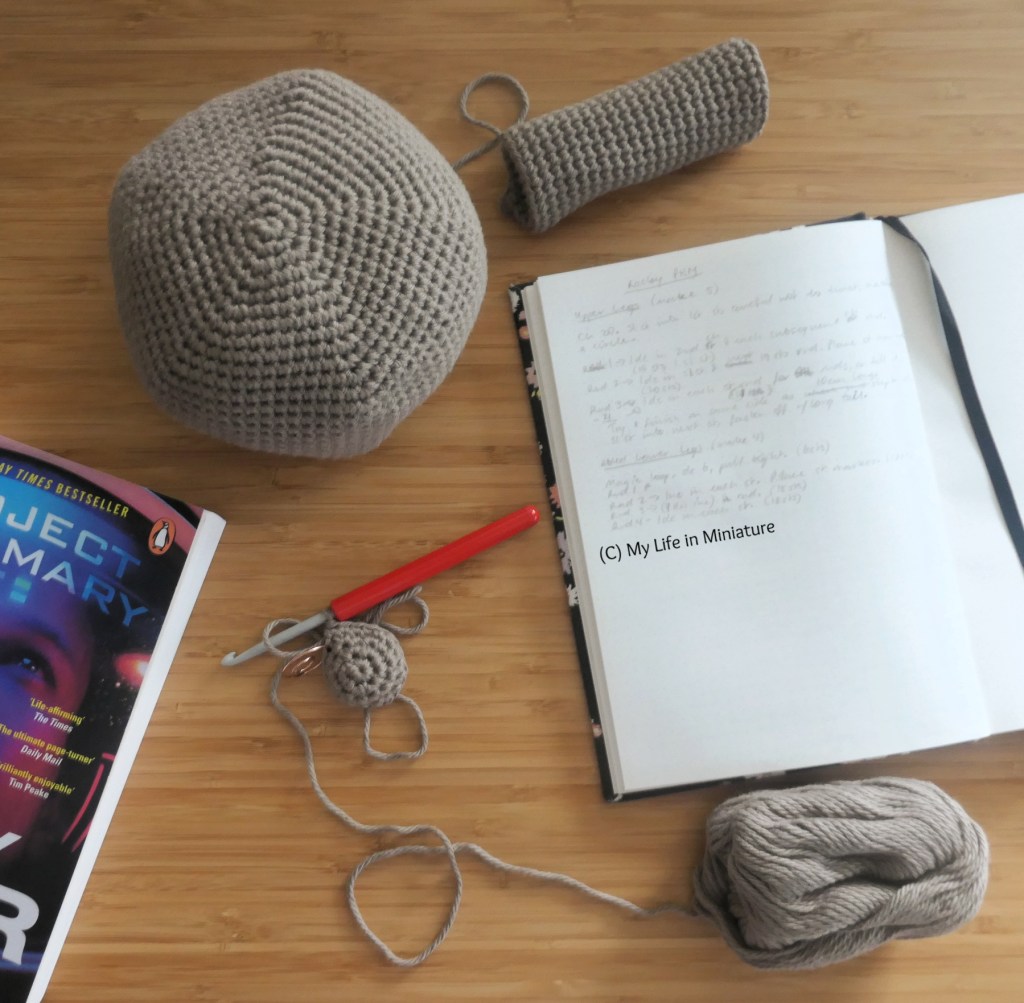 A crochet project is photographed from above on a wood surface. All three crochet pieces are made in brown yarn. Two pieces are complete, one is a tube and one is a pentagonal-ish sphere. Around the three crochet pieces are an open notebook with unreadable handwriting in, and the top corner of "Project Hail Mary" by Andy Weir.