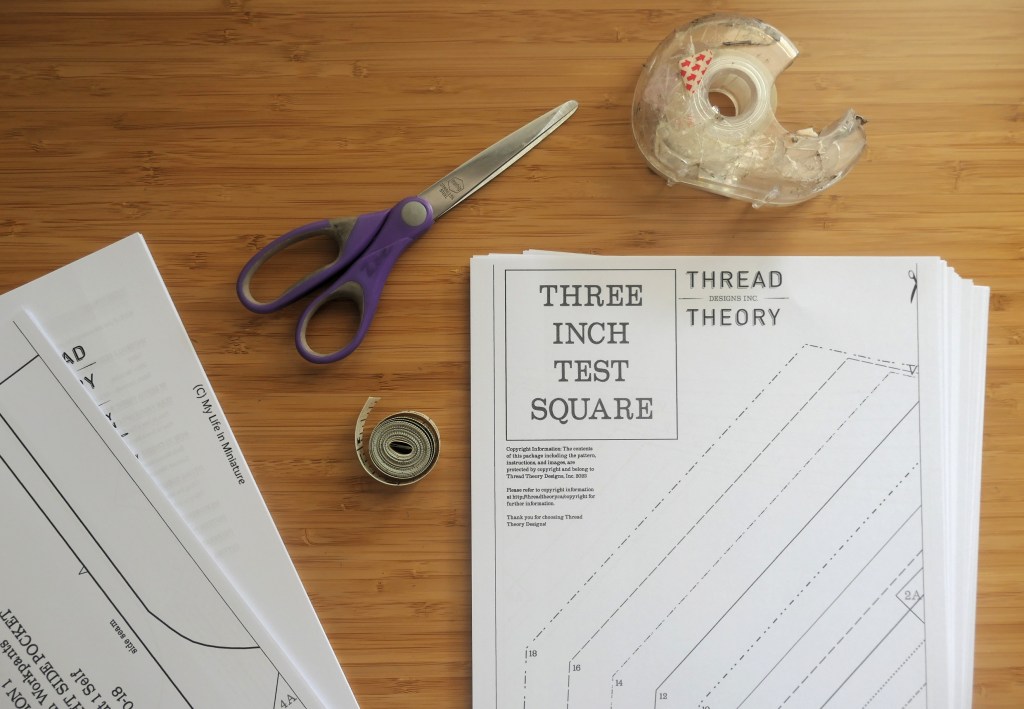 A printed sewing pattern is photographed from above on a wood surface. It is unassembled, and is in two (concerningly large) piles of paper. Around the pattern is a tape measure, a pair of paper scissors, and a sticky tape dispenser.