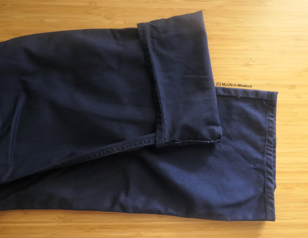 Close-up of the hems of a pair of navy pants, laid flat on a wood surface. The top leg is cuffed about 10cm, and the bottom leg is uncuffed.