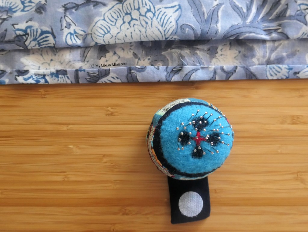Close-up of the hem of the grey floral shirt, laid flat on a wood surface. Both the back and the front sections visible have pins in them, and just below the hems is a round blue pincushion. 