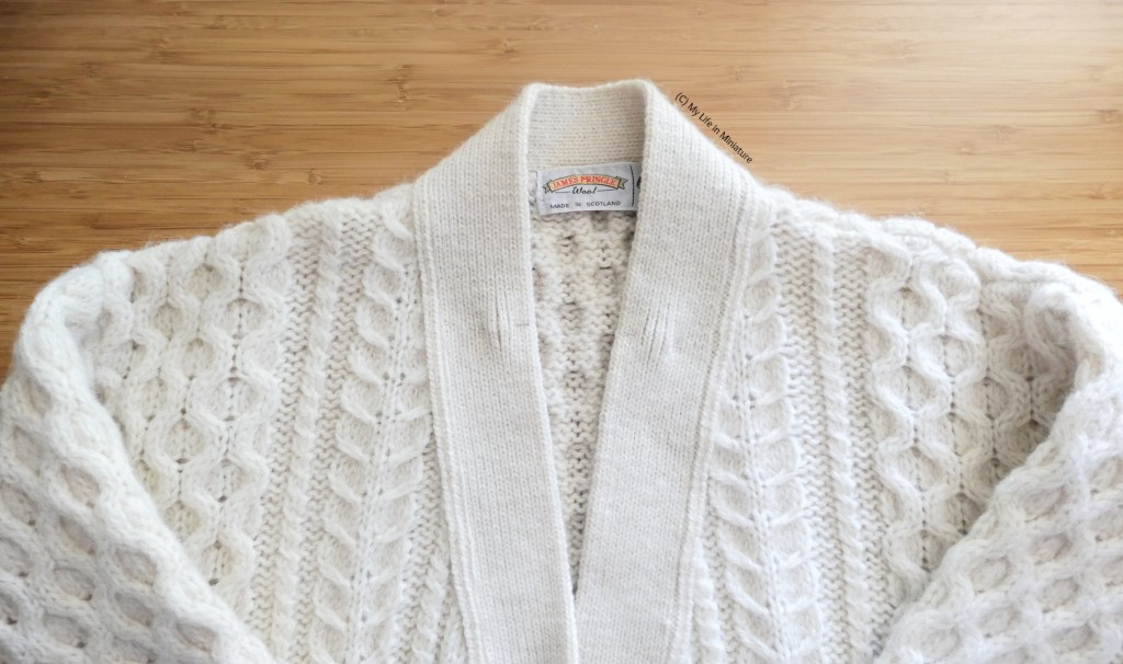 A pale beige knitted cardigan is laid flat on a wood surface, photographed from underarms up. The bodice and sleeves are heavily cabled, and the centre front opening is smooth. Thre are two bumps where pins are on the neckline, symmetrically sitting at the base of the neck.