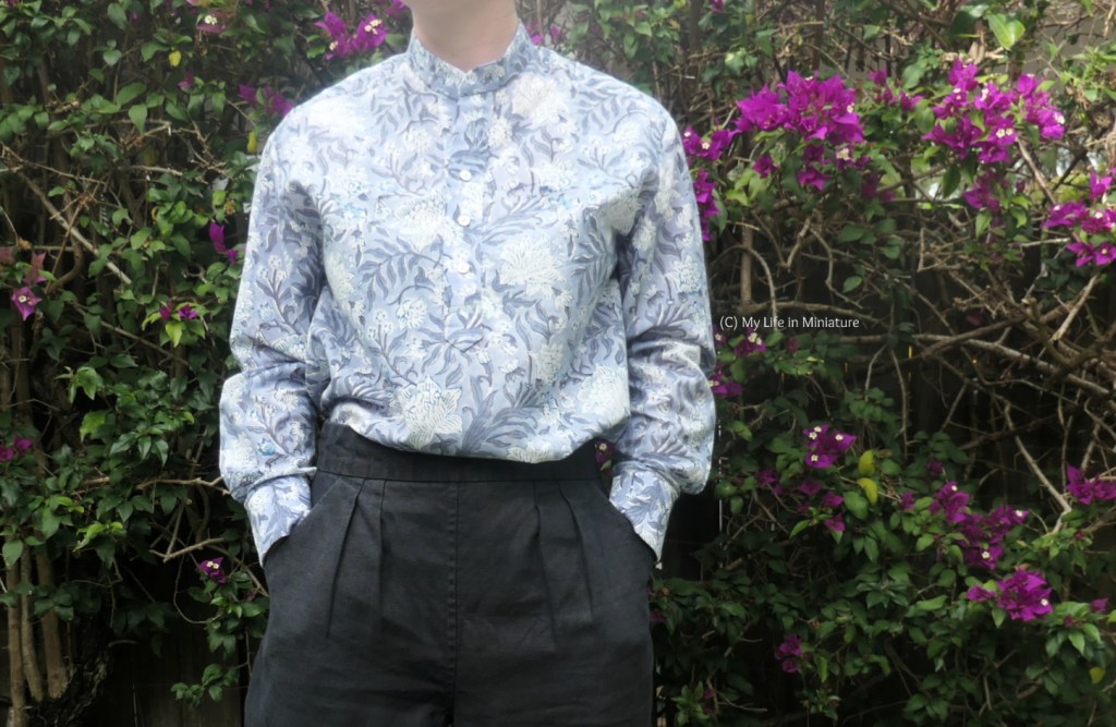 The author stands in front of a hedge, photographed from neck to thighs. She is wearing a button-down long-sleeved shirt with black pants, and has her hands in her pockets. The shirt is grey-blue, with large white flowers and darker blue stems and leaves.