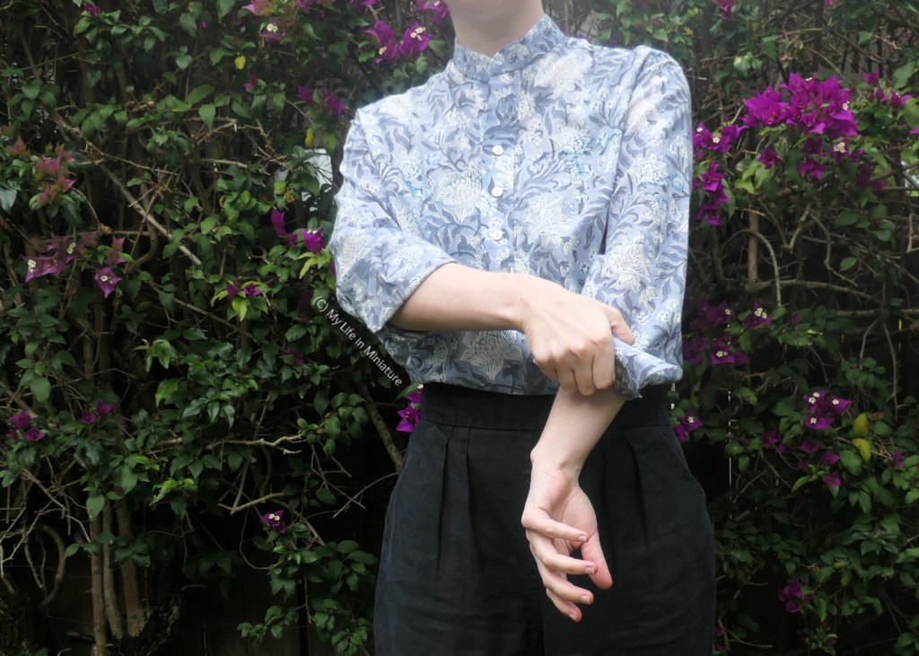 The author stands in front of a hedge, photographed from neck to thighs. She is in the middle of rolling up one of the sleeves of her shirt, having rolled the other one up already.