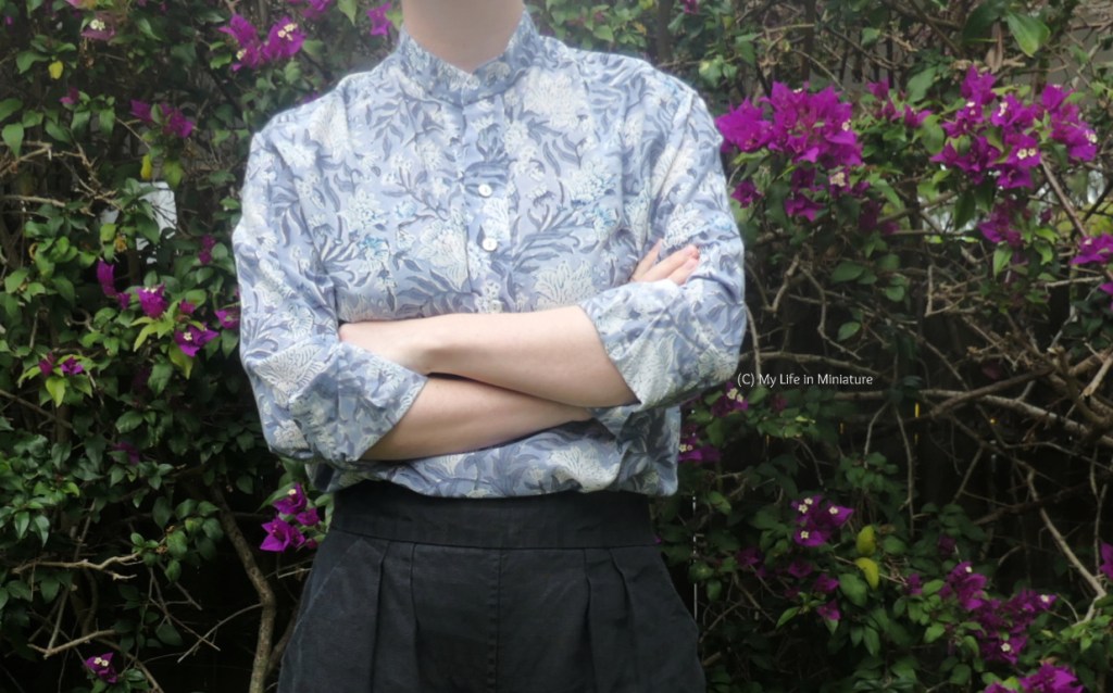 The author stands in front of a hedge, photographed from neck to thighs. She has her arms crossed, and both sleeves of her shirt rolled up. 