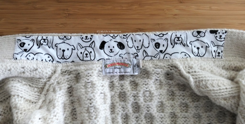 Close-up of the inside of the neck area of the knitted cardigan. A strip of white jersey printed with various dog faces in black is sewn to the inside, above the tag and past the shoulder seams.