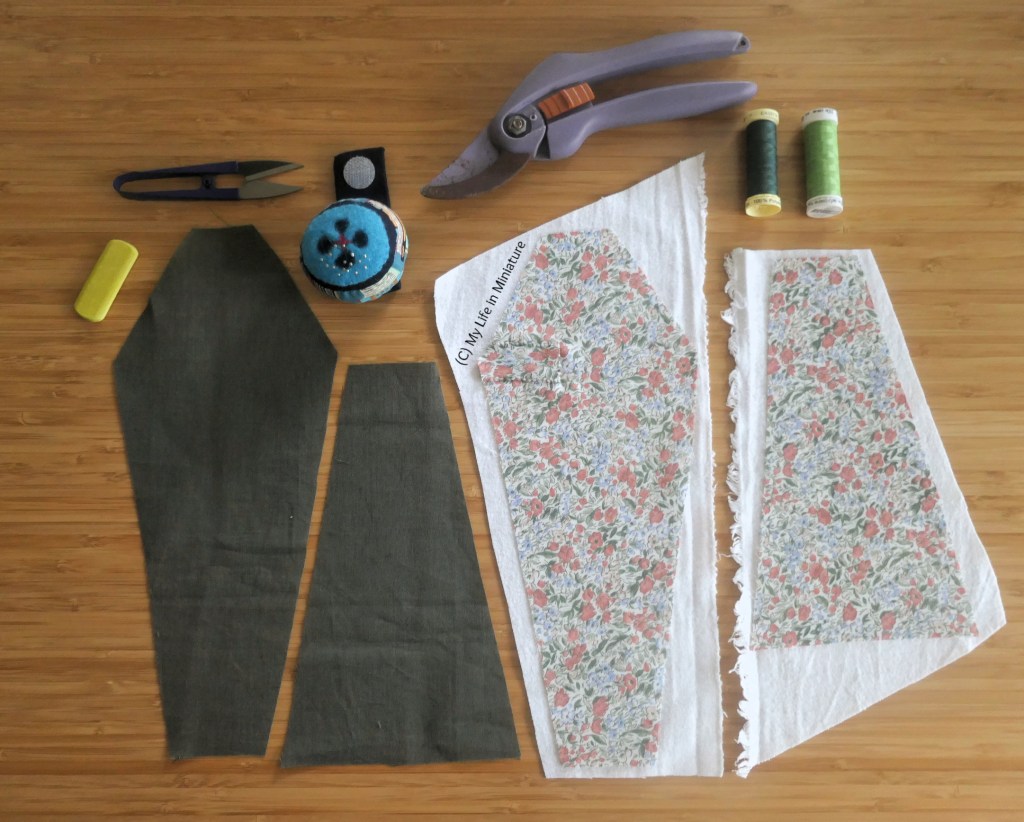 An incomplete sewing project is laid flat on a wood surface. Four pieces of fabric, two trapezoids and two elongated coffin shapes, are surrounded by various sewing notions, and a pair of secateurs. Two of the fabric shapes are dark green, and two are lighter green with red flowers.
