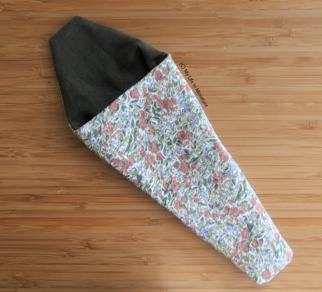 The secateur pouch is laid flat on a wood surface. It is mostly assembled; the dark green lining and floral outer are assembled and sewn together. 