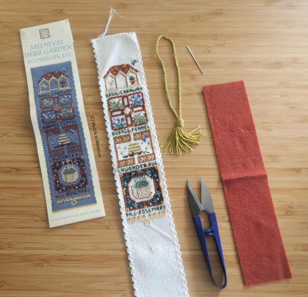 An un-assembled cross-stitch bookmark kit is laid flat on a wood surface. The paper reference image is on the left, with the completed stitching, a gold tassel, thread snips, a needle, and red backing felt alongside.