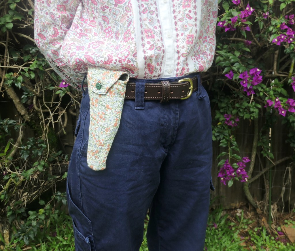 The author stands in front of a hedge, photographed from waist to knees. She's wearing the secateur pouch on the belt of her pants, which are navy. She also has a pink floral shirt tucked into her pants. The secateur pouch is buttoned shut.