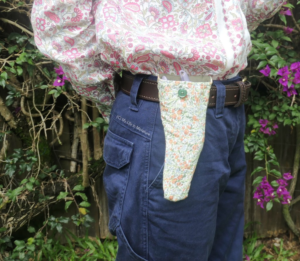 The author stands in front of a hedge, photographed from waist to knees. The secateur pouch on her belt is closer to the camera, and the flap is unbuttoned and tucked into her pants. It buttons with a plain green button, and the secateurs' grip can be seen poking out of the top.