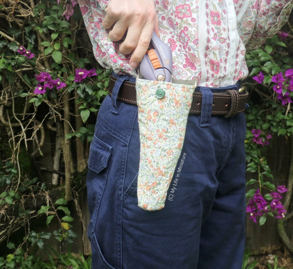 The author stands in front of a hedge, photographed from waist to knees. She holds her secateurs with one hand, holding them partially out of the pouch. The pouch is unbuttoned. 