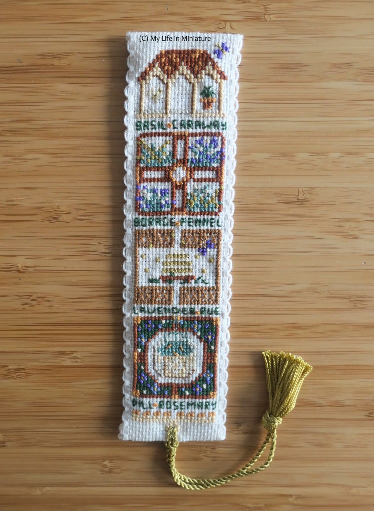 Close-up of the finished cross-stitch bookmark. It depicts a Western European medieval herb garden, with two walled gardens, a small roofed building, and a beehive. Between the small scenes is text, listing typical herbs in these gardens. The gold tassel is attached to the bottom, and curves upward.