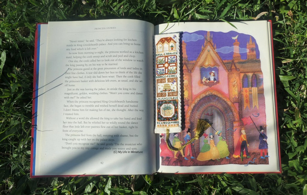 The bookmark rests in the crease of an open book, which is laid on grass in dappled sunlight. The book is a book of fairytales, with text on the left and a castle image on the right.