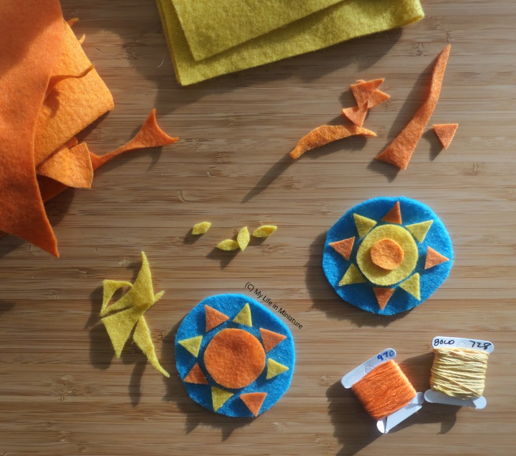 A pair of unfinished felt decorations are laid flat on a wood surface, photographed from above. The decorations are yellow and orange suns, on blue circles. Around them are scraps of yellow and orange felt, and matching embroidery thread.